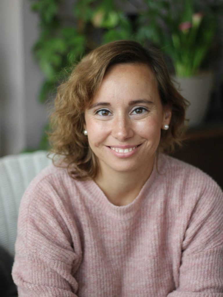 Ich bin Lara Lächelnde Frau mit lockigem Haar, gekleidet in einen rosa Pullover, vor grünen Pflanzen.